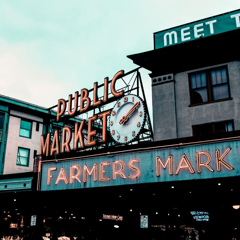 A neon sign for a public farmers market with a clock is displayed on a building, beneath a secondary sign that reads "MEET THE."