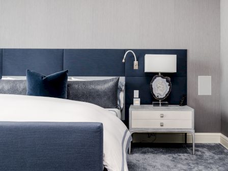 A modern bedroom with a navy blue upholstered bed, white linens, bedside table with a lamp and decorative object, and gray carpet.