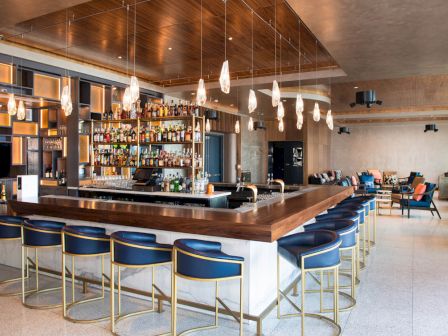 This image shows a modern bar with a well-stocked back wall, blue bar stools, pendant lighting, and seating areas in a stylish interior space.