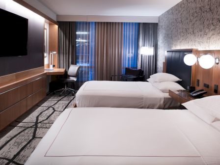 A modern hotel room with two double beds, a large flat-screen TV, a desk with a chair, bedside lamps, and floor-to-ceiling windows with curtains.
