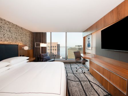 A modern hotel room with a large bed, a desk and chair, a flat-screen TV, and a window with a city view.
