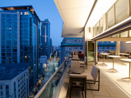 The image shows a modern urban balcony overlooking a city street with high-rise buildings, open glass doors, and seating arrangements on the patio.