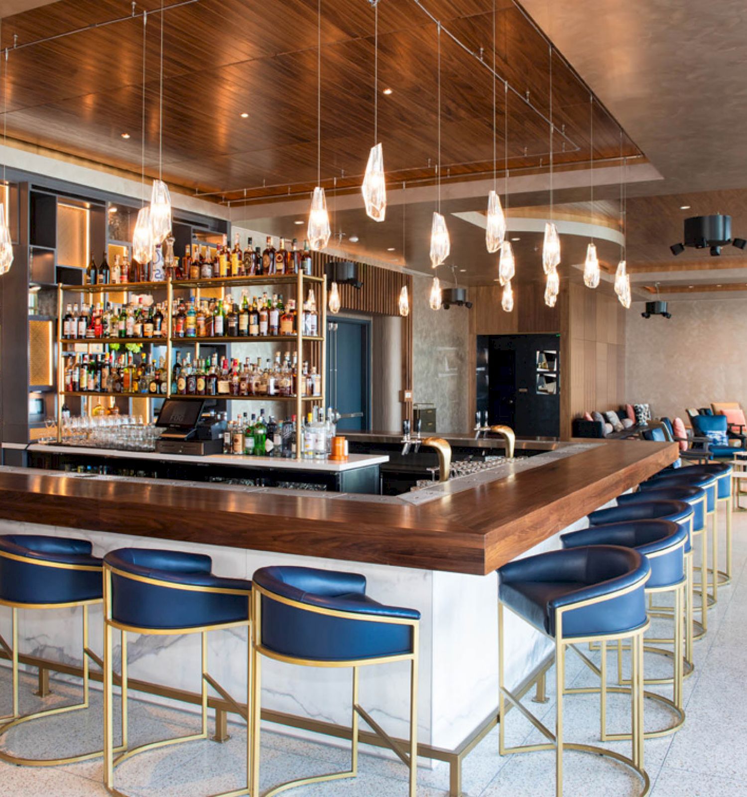 The image shows a modern bar with a central counter, barstools, hanging lights, and a well-stocked liquor display. It has seating arrangements in the background.