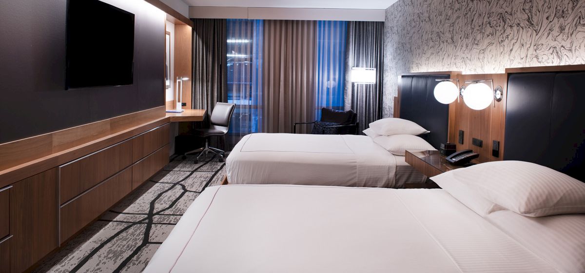 A modern hotel room with two beds, a wall-mounted TV, desk, chair, lamps, and floor-to-ceiling windows with long curtains ending the sentence.