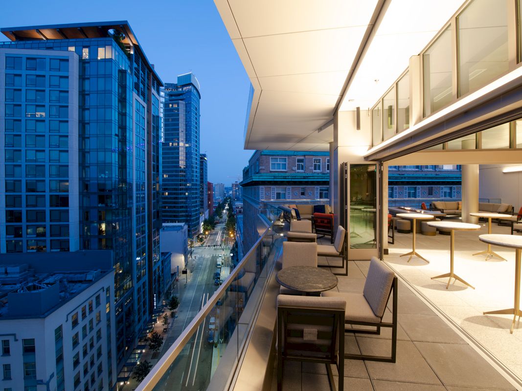 The image shows a modern city view with tall buildings, streets, and a stylish balcony with tables and chairs at dusk.