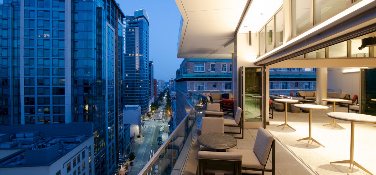 The image shows a modern city view with tall buildings, streets, and a stylish balcony with tables and chairs at dusk.