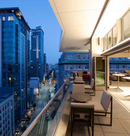 The image shows a rooftop terrace with seating and tables overlooking a cityscape with tall buildings illuminated during twilight hours.