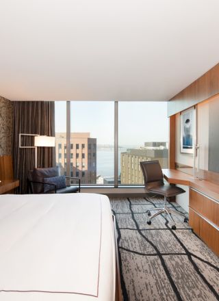 The image shows a modern hotel room with a large bed, patterned wallpaper, a flat-screen TV, a desk, a chair, and a view of buildings through a window.