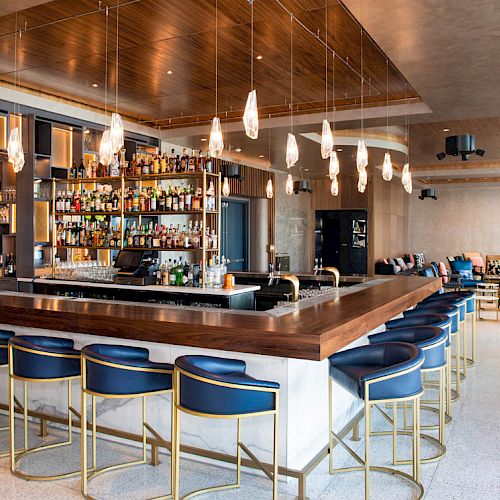 This image shows a modern bar with blue stools, a large selection of drinks behind the counter, and contemporary pendant lights hanging from the ceiling.