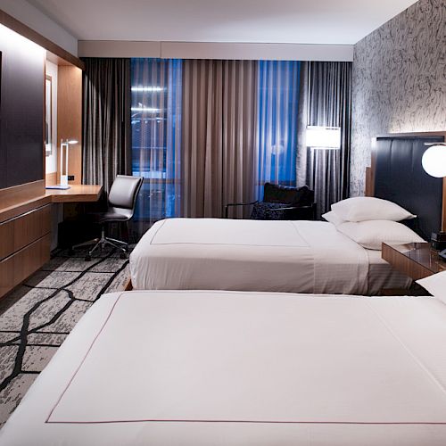 The image shows a modern hotel room with two beds, a wall-mounted TV, desk and chair, bedside tables, patterned carpet, and large windows with curtains.
