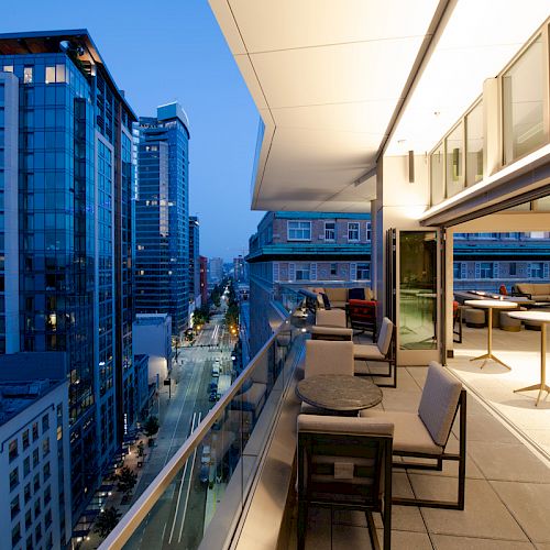 The image shows a modern urban balcony at dusk with seating, a glass railing, and city buildings with lights and skyscrapers in the background.