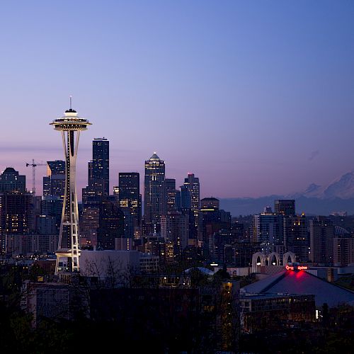 A skyline with the Space Needle, various high-rises, and a distant mountain under a twilight sky, lit up urban and natural scenery combined.