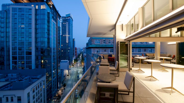 A modern balcony view overlooking a city street at dusk, with seating areas and tables, adjacent to tall buildings with large glass windows.