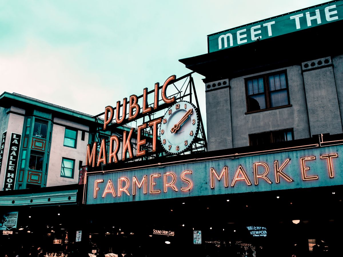 The image features a neon sign for a "Public Market" with a clock and a "Farmers Market" sign below, set against urban buildings.