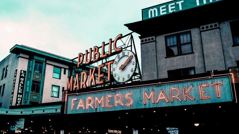 The image features a neon sign for a "Public Market" with a clock and a "Farmers Market" sign below, set against urban buildings.