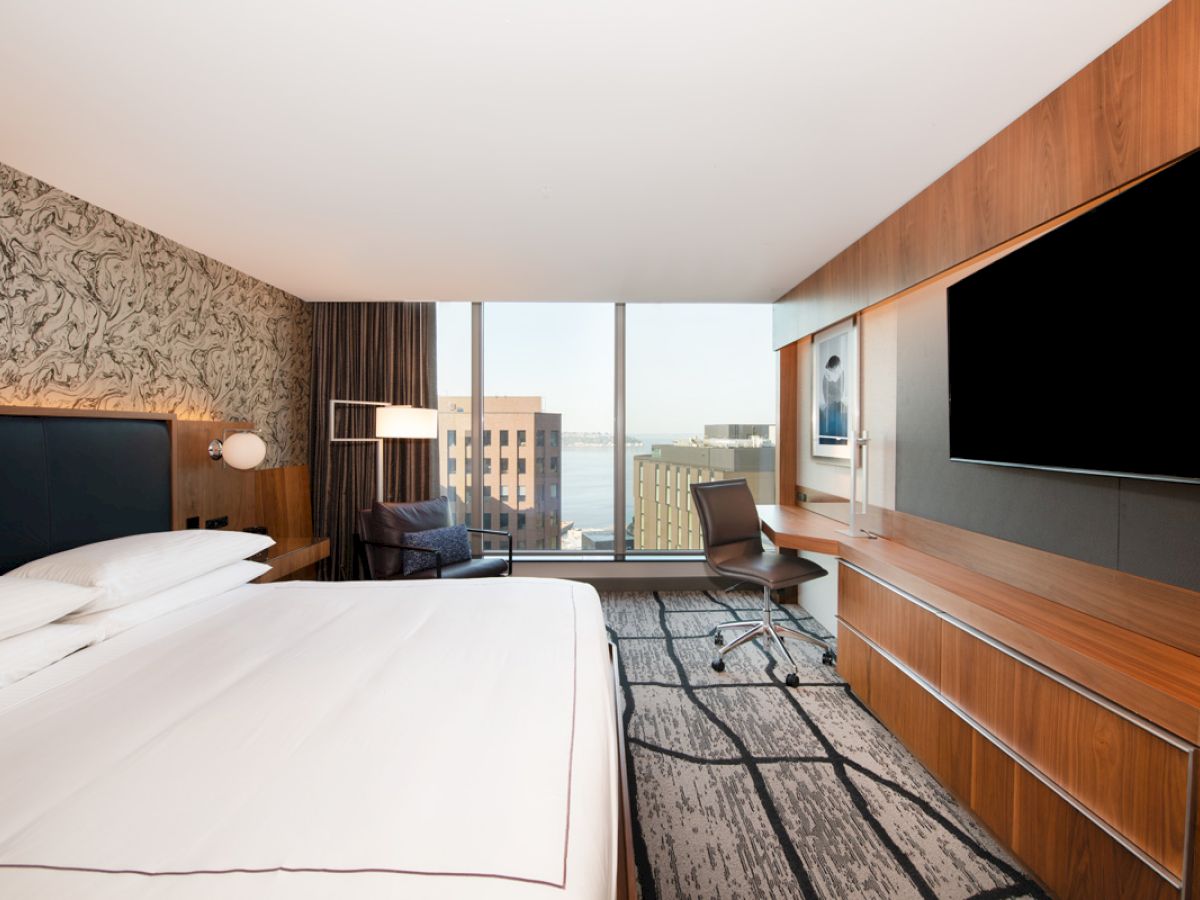 A modern hotel room with a large bed, wall-mounted TV, desk, chair, and large window with a city view, featuring contemporary decor.