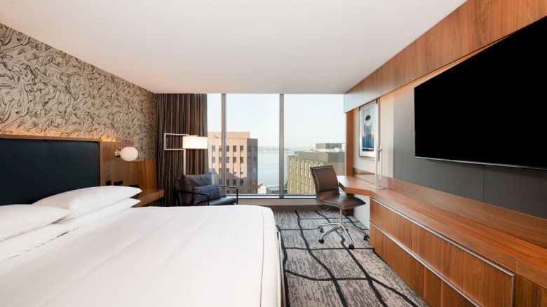 A modern hotel room with a large bed, wall-mounted TV, desk, chair, and large window with a city view, featuring contemporary decor.
