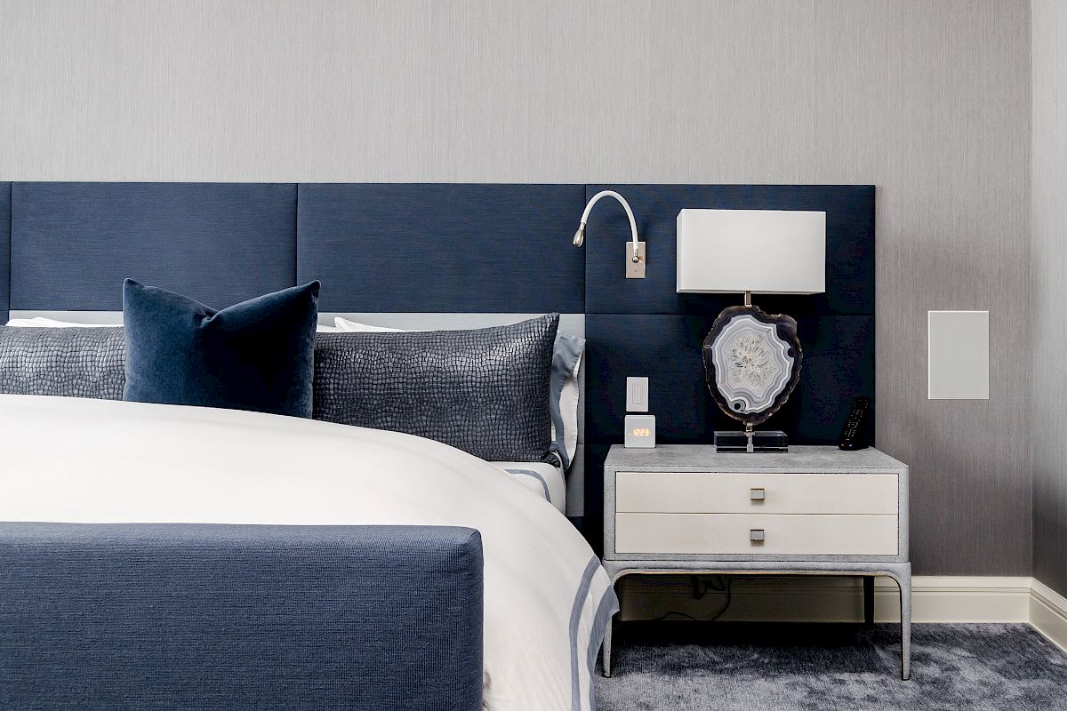 A bedroom with a blue and white color scheme, featuring a bed with blue headboard, nightstand with a lamp and decor, and gray carpet.