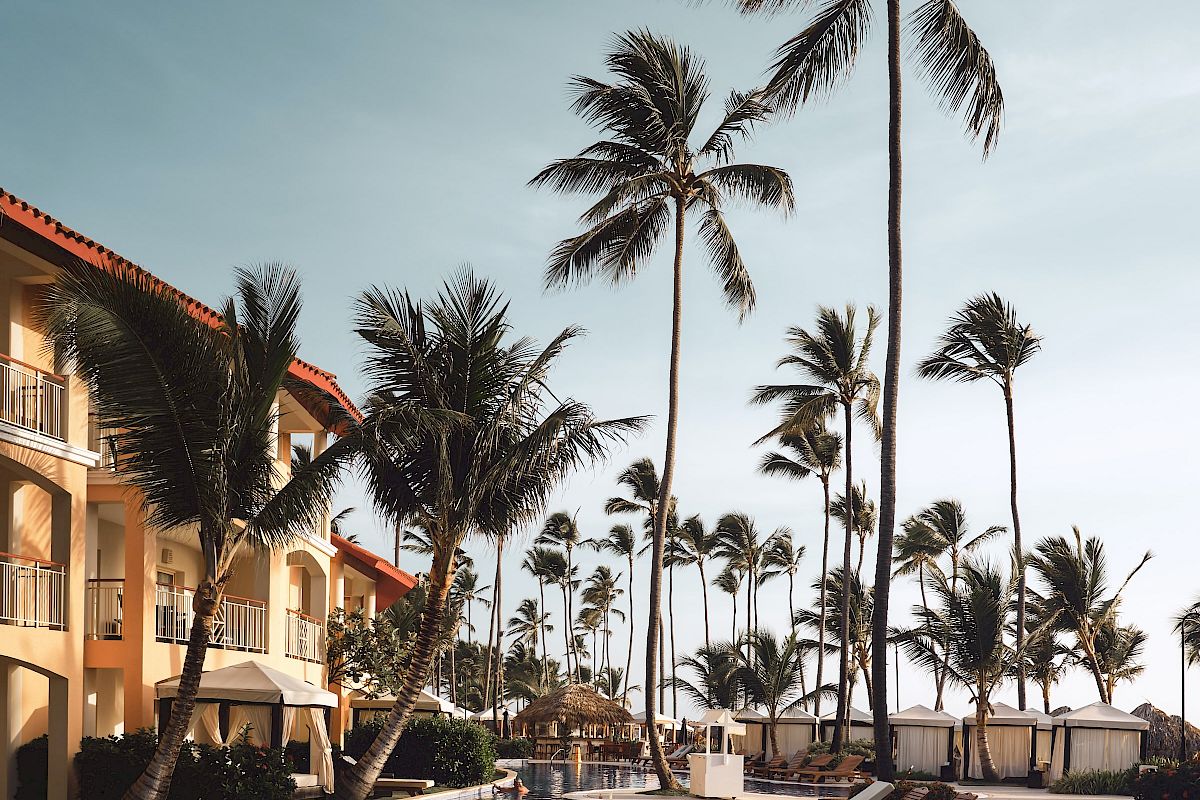 The image shows a tropical resort with a serene pool, surrounded by deck chairs, palm trees, and adjacent buildings under a clear sky.