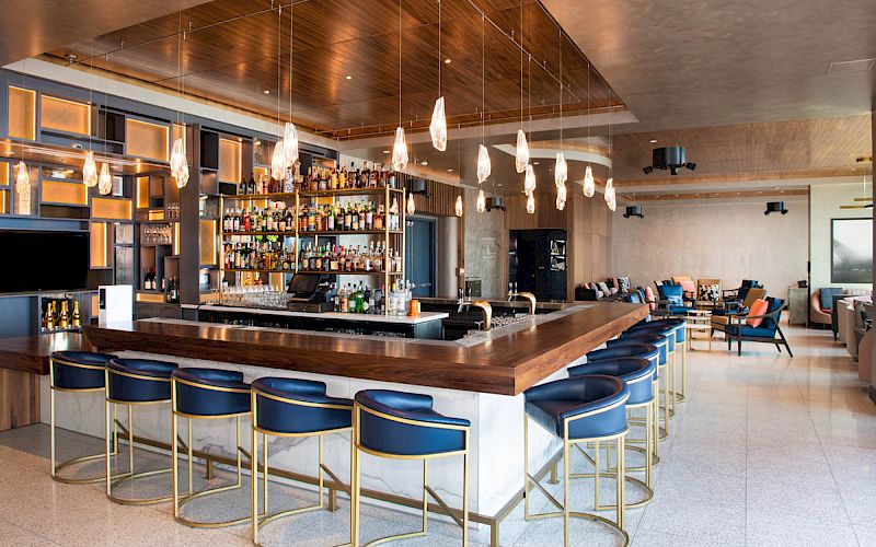 The image shows a modern bar with a wooden counter, blue bar stools, hanging lights, and a well-stocked shelf of liquor bottles in a sleek interior setting.