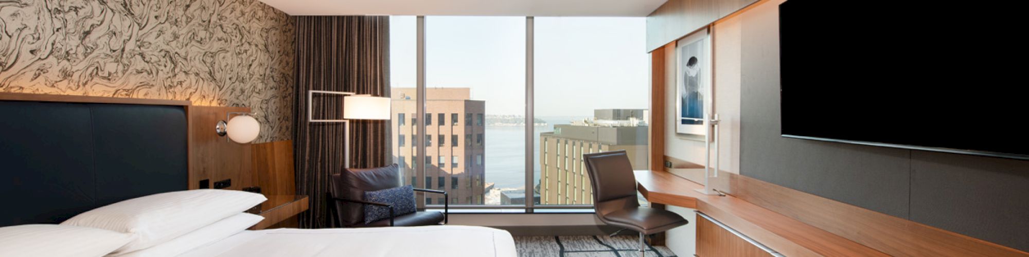 A modern hotel room with a large bed, wall-mounted TV, desk, chair, and large window offering a cityscape view.